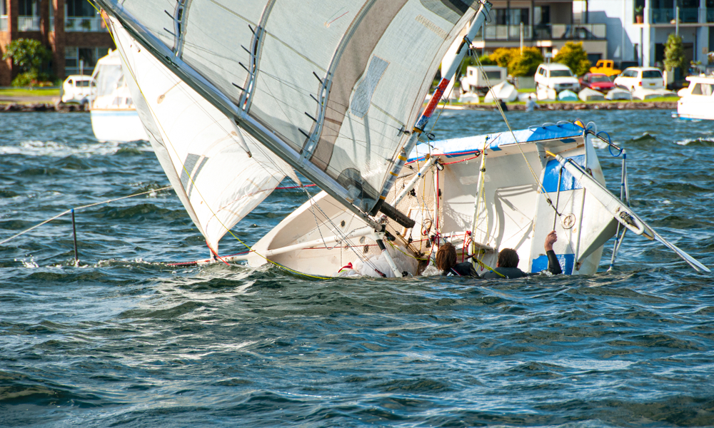 sinking sailboat near a port turned on its side while the people in the water try to get get back to the boat