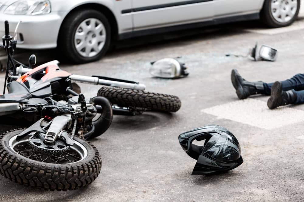 Scene of a motorcycle accident with a motorcycle lying on its side on the road. A helmet is on the ground nearby, and a person is lying on the pavement. Broken car parts are scattered around, with a car visible in the background.