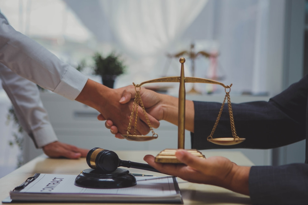 close up shot of two people shaking hands across a desktop which has the scales of justice and a judge's gavel atop it