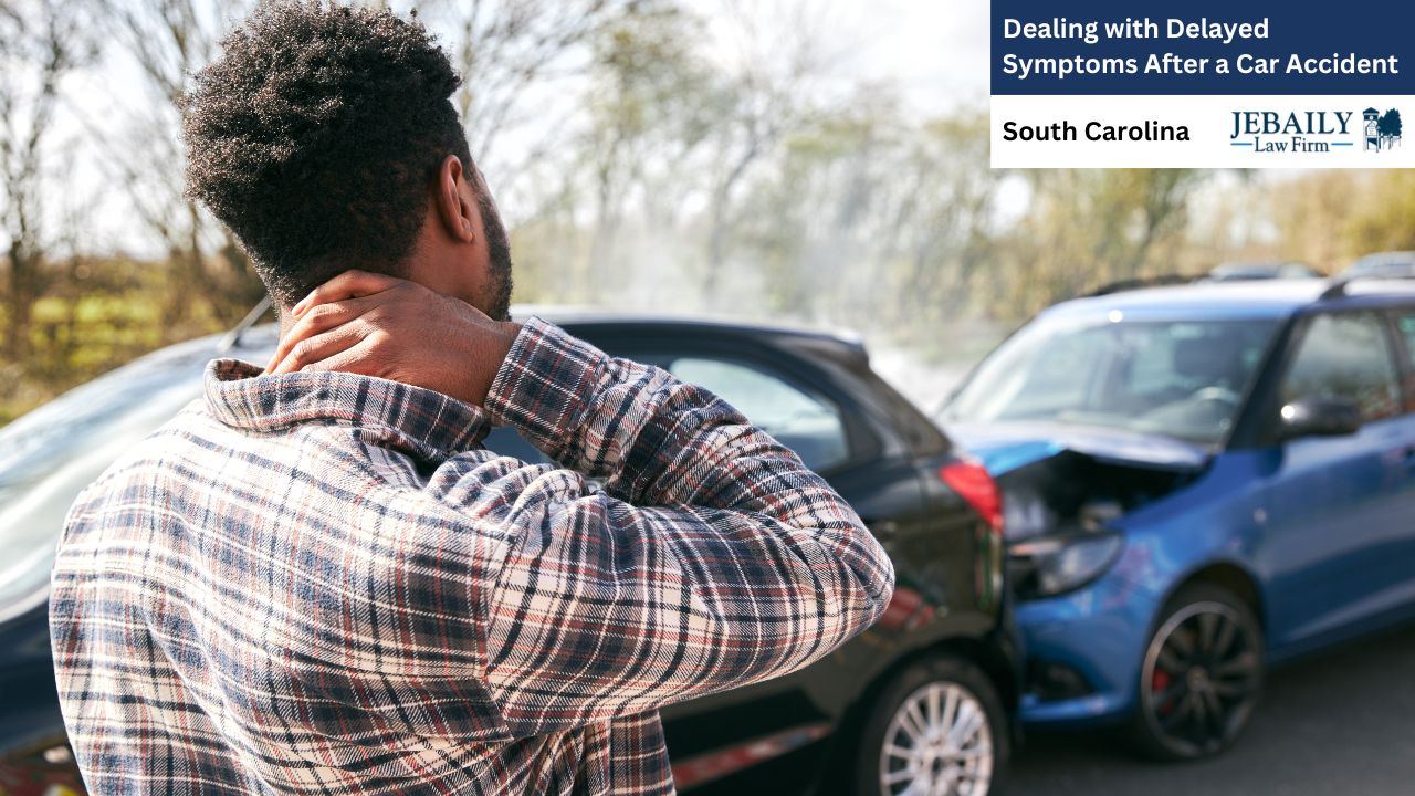 A man is experiencing delayed symptoms after a car accident. The image shows a Black man from behind, wearing a plaid shirt, who is holding his neck with both hands, appearing to be in pain. His focus is inward, suggesting discomfort or distress. In the background, out of focus but clearly visible, are two damaged cars.