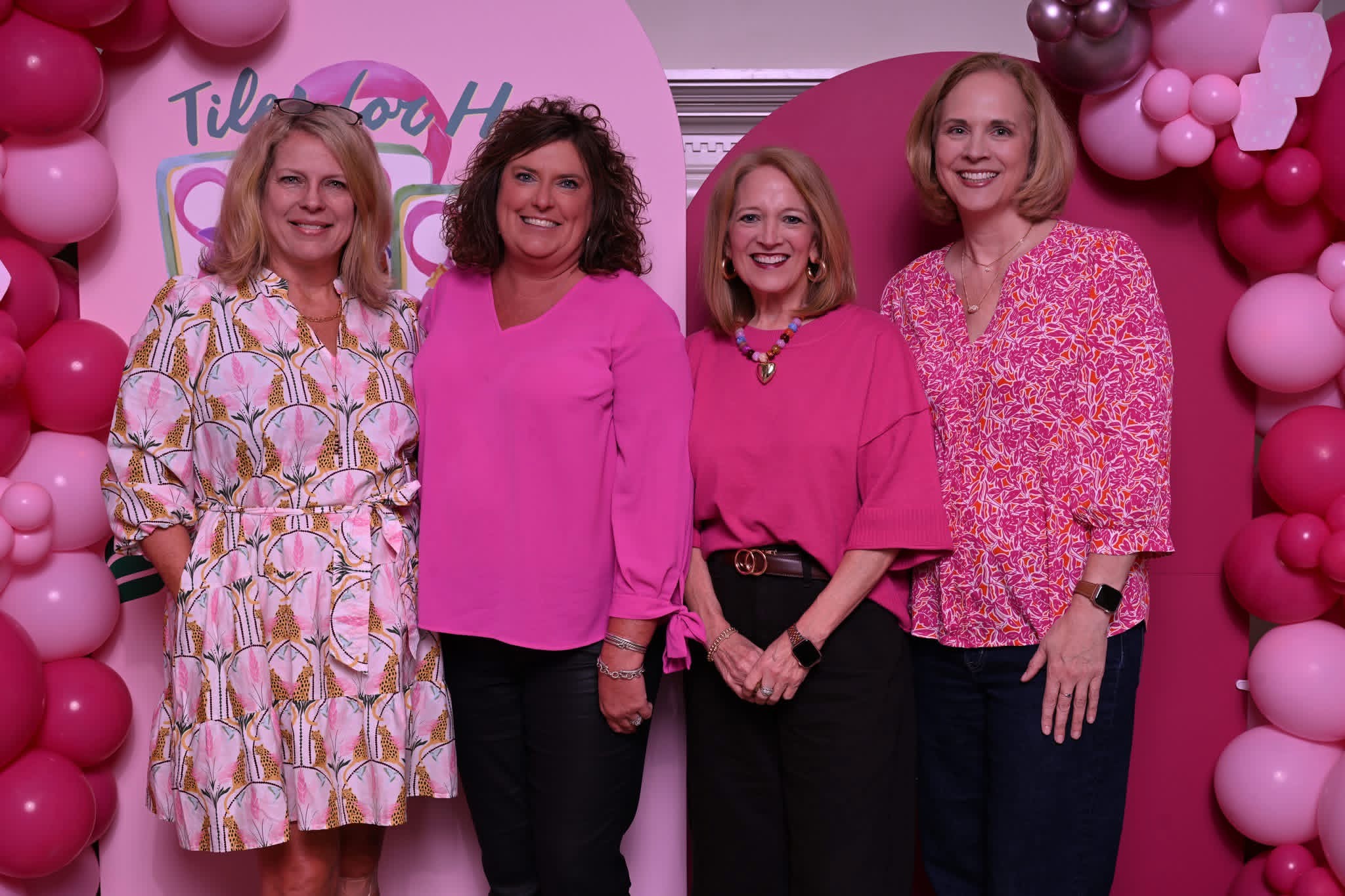 Four women wearing variations of pink tops and dresses with black pants stand in front of a pink Tiles of Hope sign.